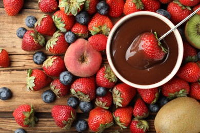 Photo of Fondue fork with strawberry in bowl of melted chocolate surrounded by other fruits on wooden table, flat lay