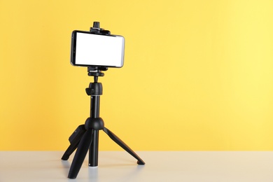Photo of Smartphone with blank screen fixed to tripod on white table against yellow background. Space for text