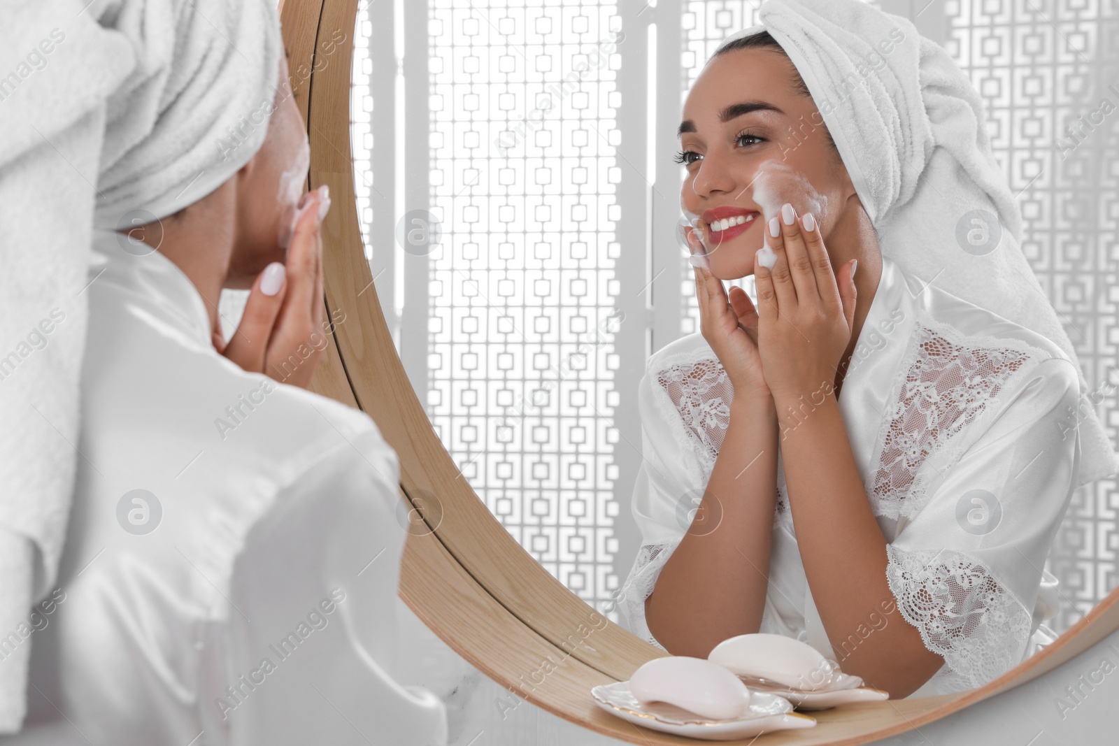 Photo of Beautiful young woman applying cleansing foam onto face near mirror in bathroom. Skin care cosmetic