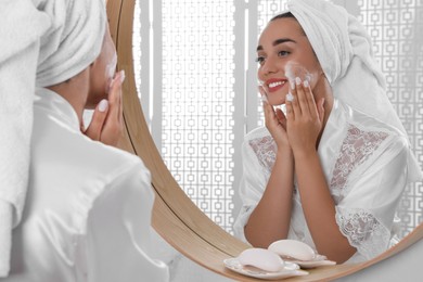 Photo of Beautiful young woman applying cleansing foam onto face near mirror in bathroom. Skin care cosmetic