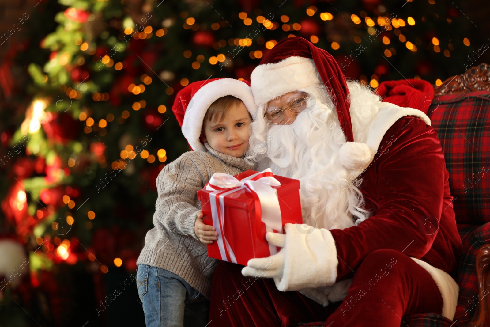 Photo of Santa Claus and little boy with gift near Christmas tree indoors