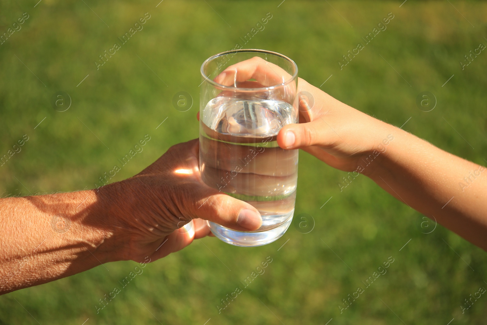 Photo of Child giving glass of water to elderly woman outdoors on sunny day, closeup