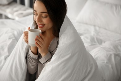 Photo of Beautiful young woman wrapped with soft blanket holding cup of hot drink on bed at home