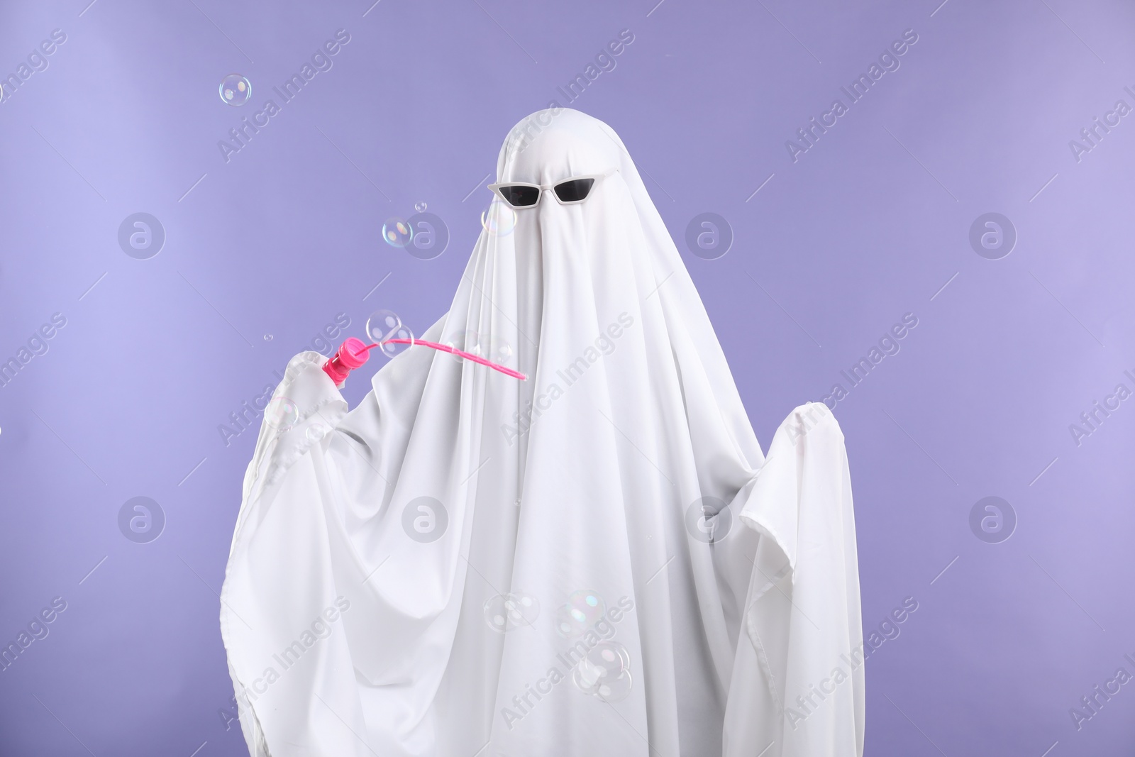 Photo of Funny ghost. Person in white sheet and sunglasses blowing soap bubbles on violet background