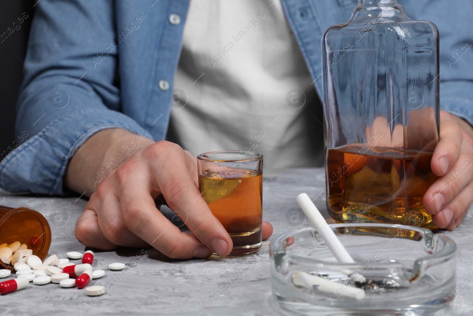 Photo of Alcohol and drug addiction. Man with whiskey, pills and smoldering cigarettes at grey textured table, closeup