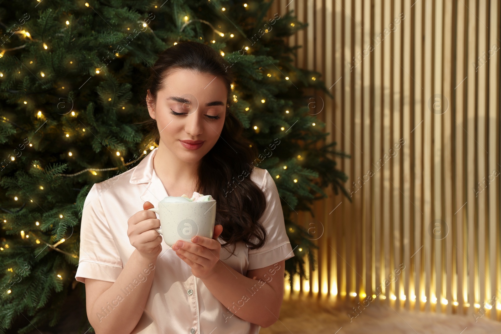 Photo of Beautiful woman with cup of tasty drink near Christmas tree indoors