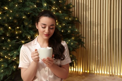 Beautiful woman with cup of tasty drink near Christmas tree indoors