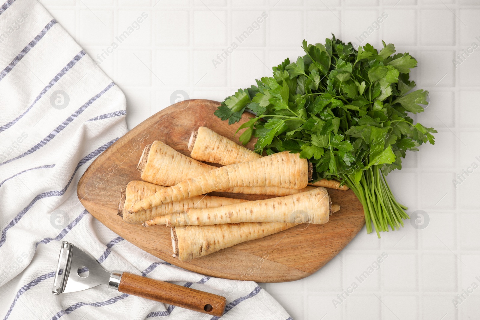 Photo of Raw parsley roots, bunch of fresh herb and peeler on white table, flat lay