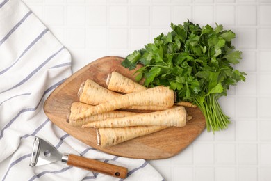 Raw parsley roots, bunch of fresh herb and peeler on white table, flat lay