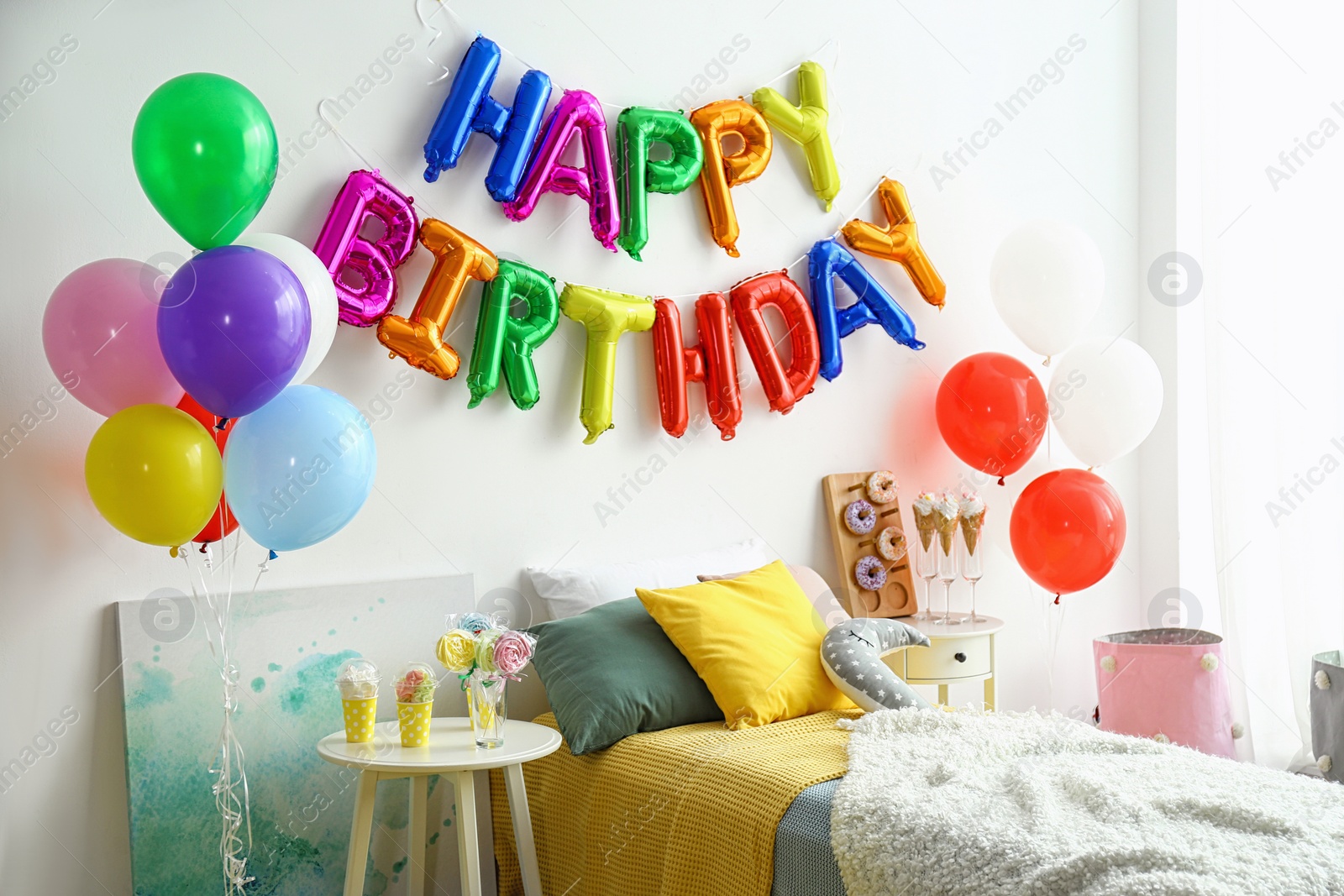 Photo of Phrase HAPPY BIRTHDAY made of colorful balloon letters and table with treats in modern bedroom