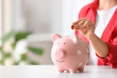 Photo of Woman putting coin into piggy bank at table indoors, closeup. Space for text