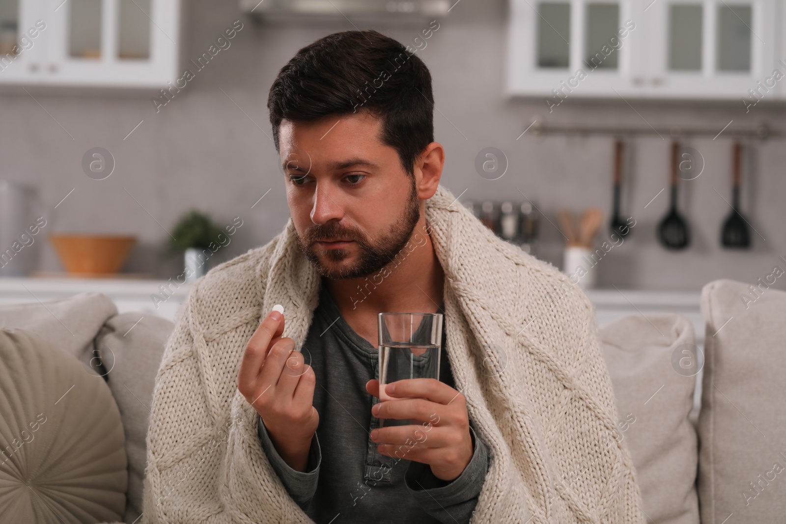 Photo of Depressed man holding glass of water and antidepressant pill on sofa at home