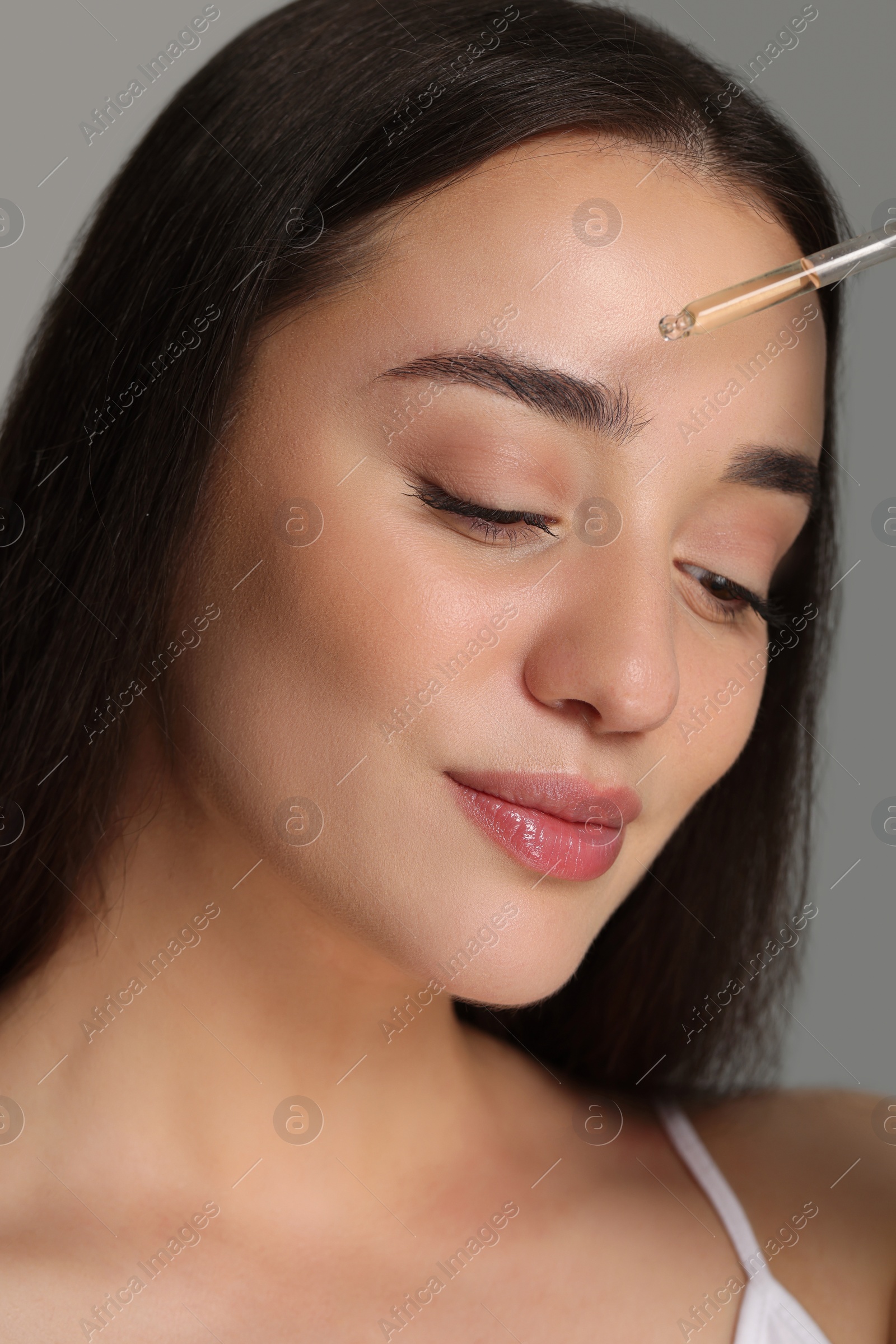 Photo of Beautiful young woman applying essential oil onto face on grey background, closeup