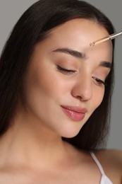 Beautiful young woman applying essential oil onto face on grey background, closeup