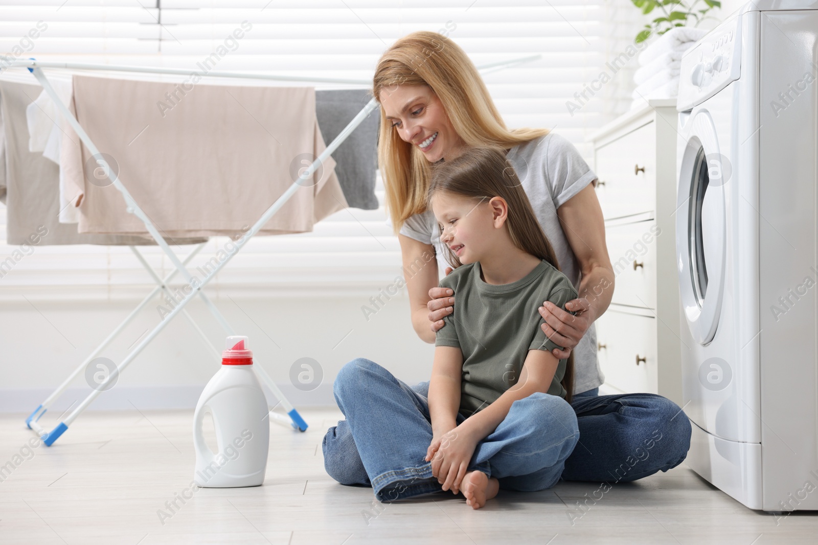 Photo of Mother and daughter sitting on floor near washing machine and looking at fabric softener in bathroom, space for text