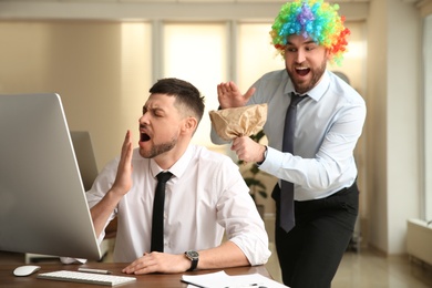 Photo of Young man popping paper bag behind his colleague in office. Funny joke