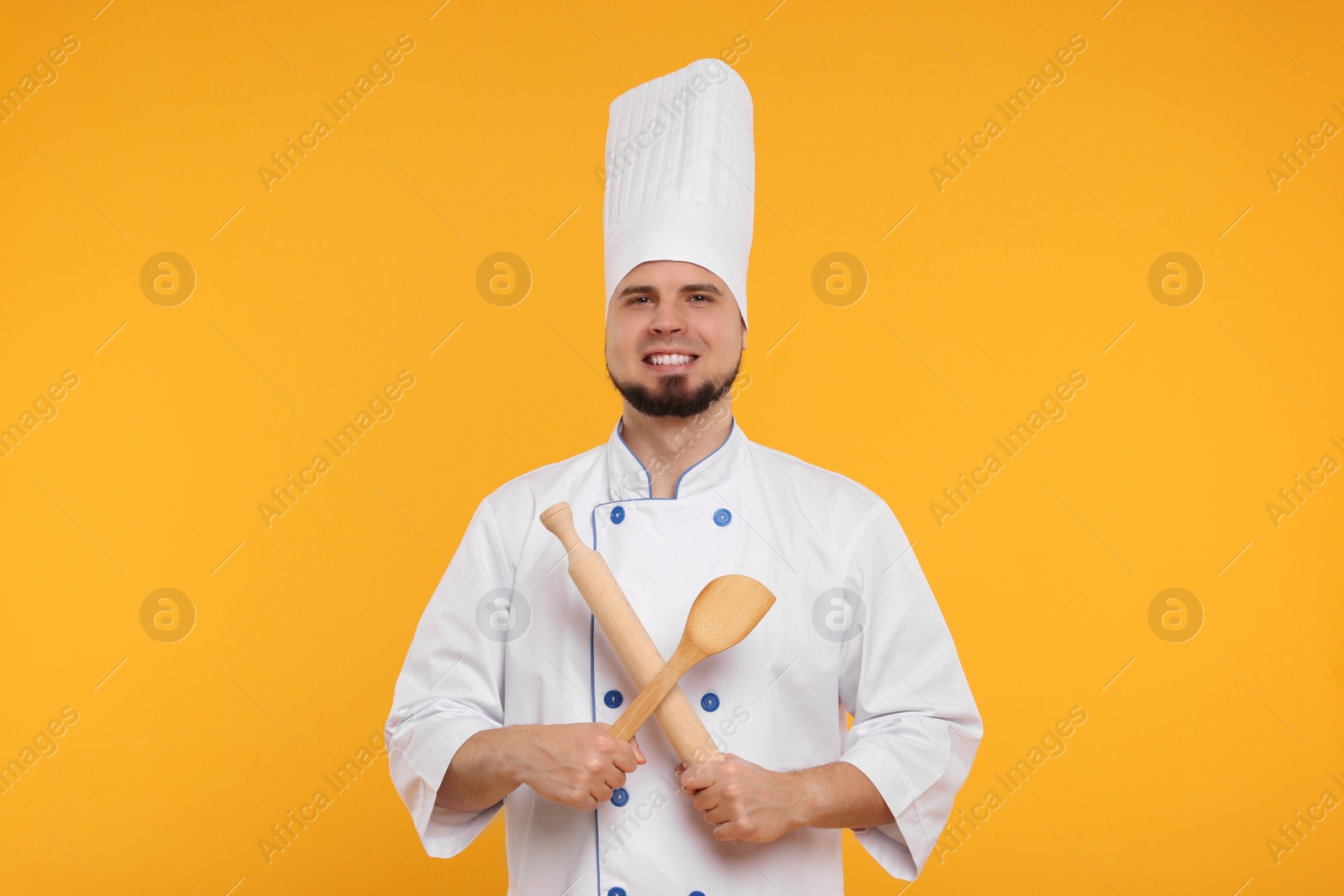 Photo of Happy professional confectioner in uniform holding rolling pin and spatula on yellow background