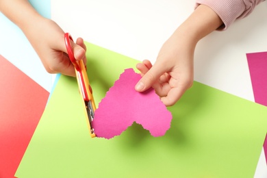 Photo of Child cutting out paper heart with craft scissors at table, top view