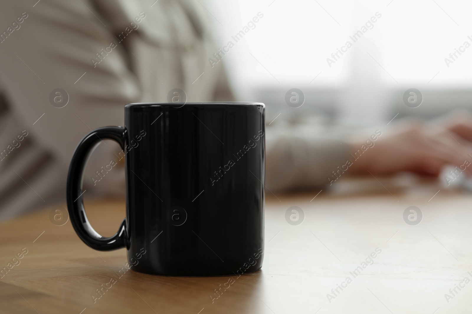 Photo of Black ceramic mug on wooden table. Woman at workplace, selective focus. Space for text