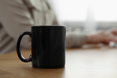 Black ceramic mug on wooden table. Woman at workplace, selective focus. Space for text