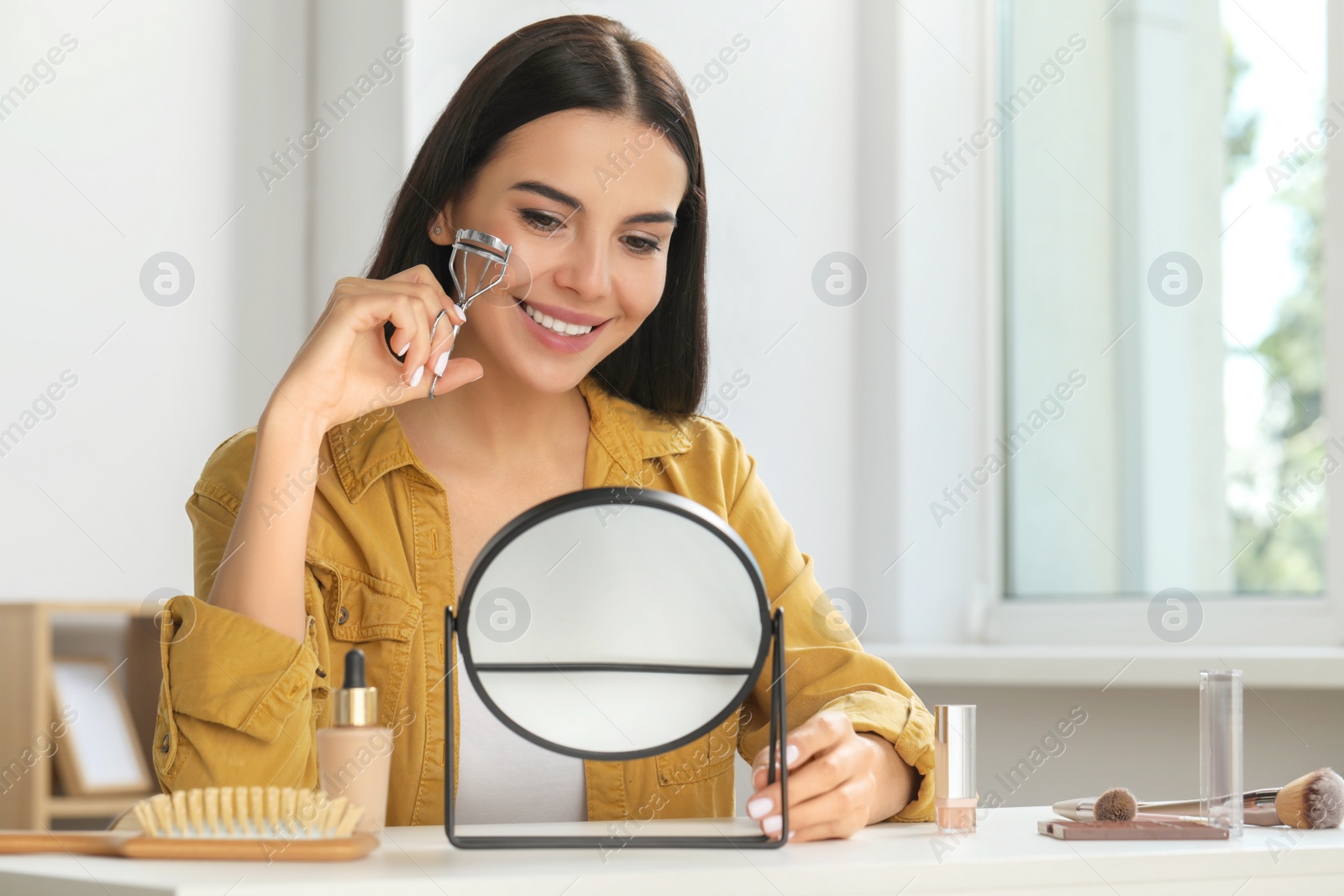 Photo of Beautiful young woman using eyelash curler while doing makeup at table indoors