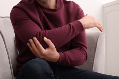 Man suffering from pain in his elbow on armchair indoors, closeup