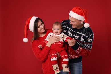 Photo of Happy couple with cute baby in Christmas outfits and Santa hats on red background
