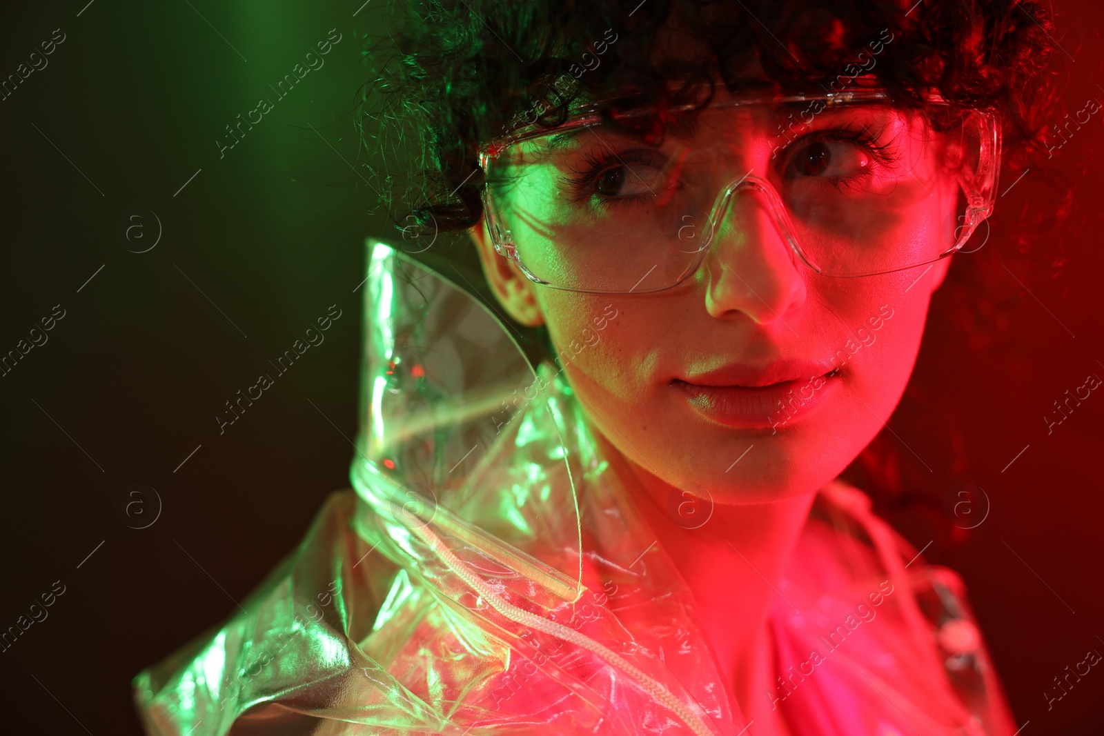 Photo of Beautiful young woman in transparent coat and sunglasses posing on color background in neon lights, closeup