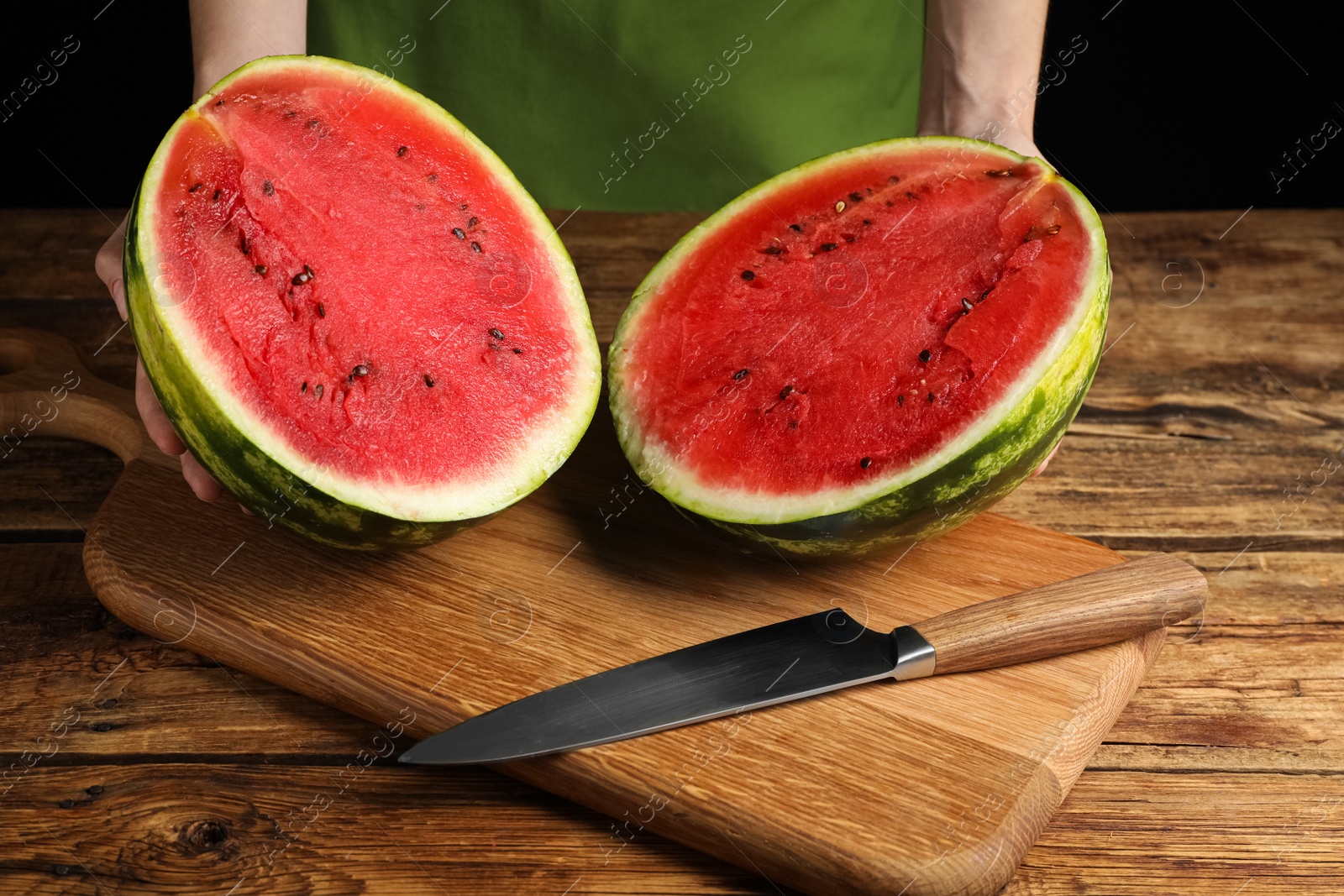 Photo of Woman with delicious halved watermelon at wooden table against black background, closeup