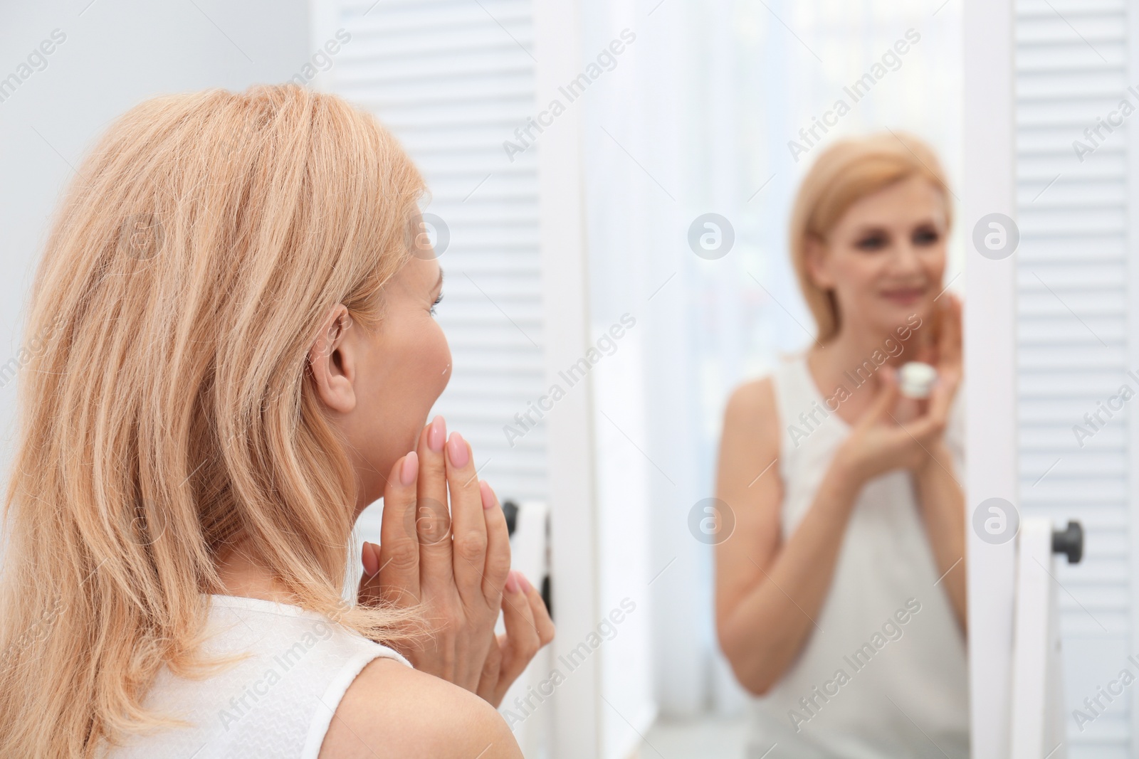 Photo of Mature woman applying face cream near mirror indoors. Space for text