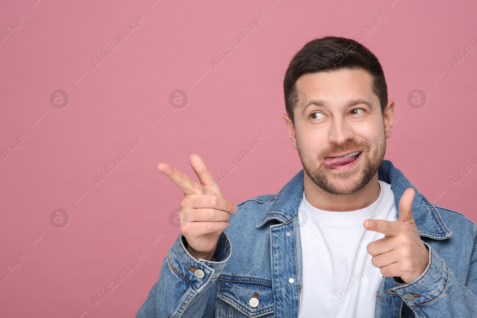 Photo of Happy man showing his tongue and pointing at something on pink background. Space for text