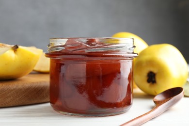 Photo of Tasty homemade quince jam in jar, spoon and fruits on white wooden table, closeup