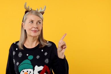 Photo of Senior woman in Christmas sweater and deer headband pointing at something on orange background. Space for text