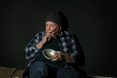Photo of Poor senior man with bread and bowl on floor near dark wall