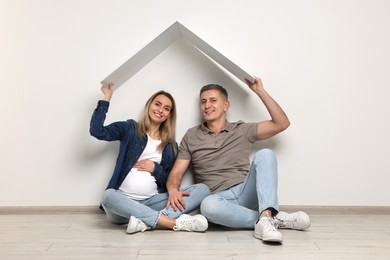 Young family housing concept. Pregnant woman with her husband sitting under cardboard roof on floor indoors
