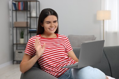 Happy young woman having video chat via laptop and waving hello on sofa in living room