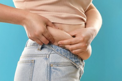 Woman touching belly fat on light blue background, closeup. Overweight problem