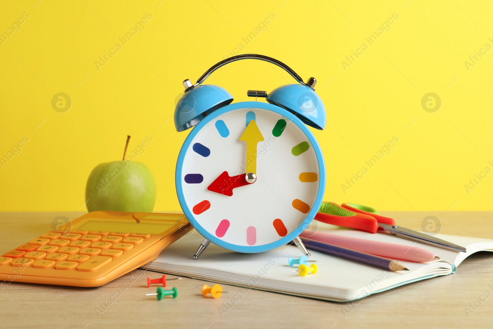 Photo of Light blue alarm clock and different stationery on white wooden table against yellow background. School time