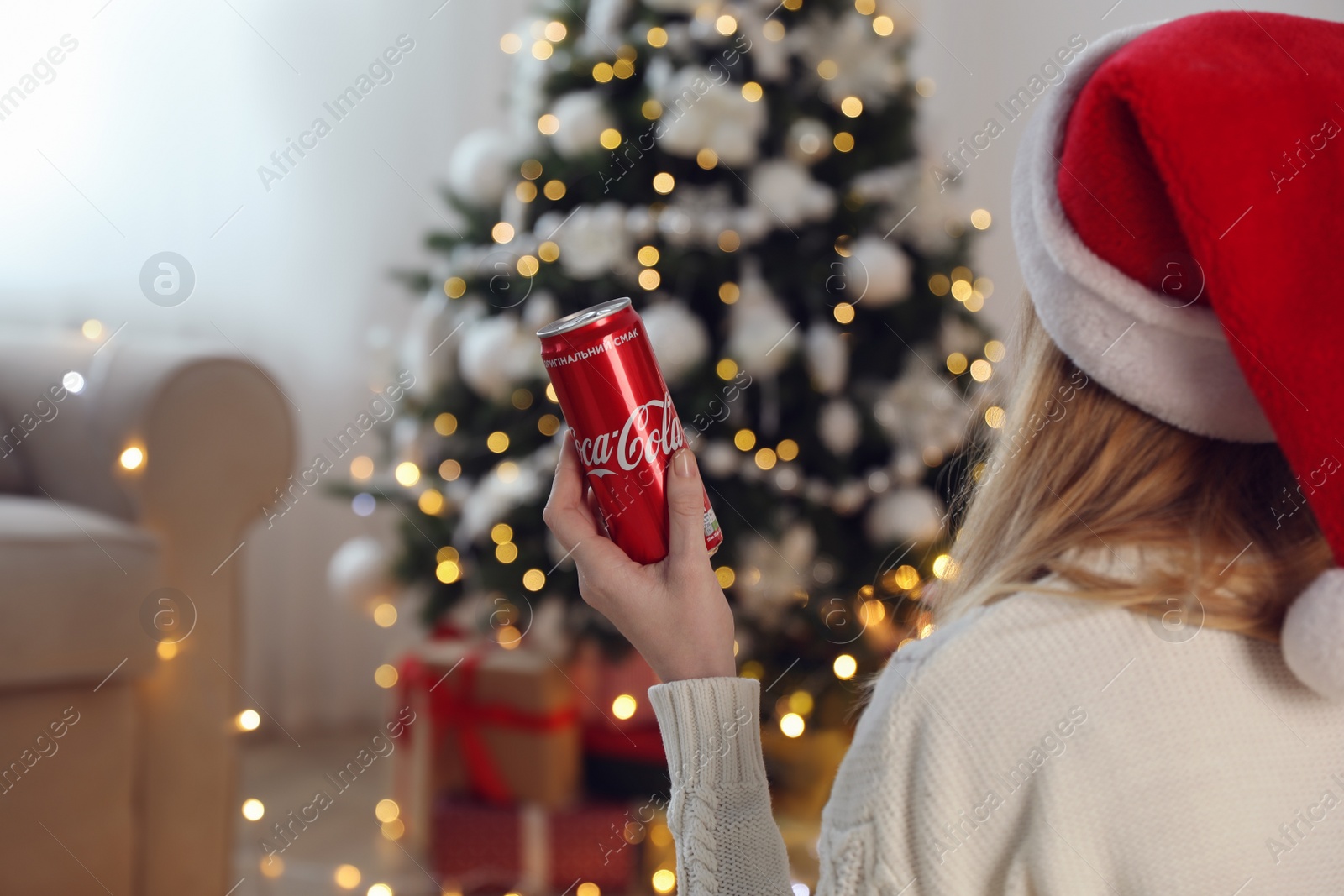 Photo of MYKOLAIV, UKRAINE - January 01, 2021: Woman in Santa hat with can of Coca-Cola against blurred Christmas tree at home, closeup