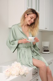 Photo of Pretty young woman in beautiful silk robe with cup of coffee sitting on kitchen counter