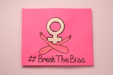 Photo of Card with hashtag BreakTheBias, gender female symbol and drawing of crossed arms on pink background, top view