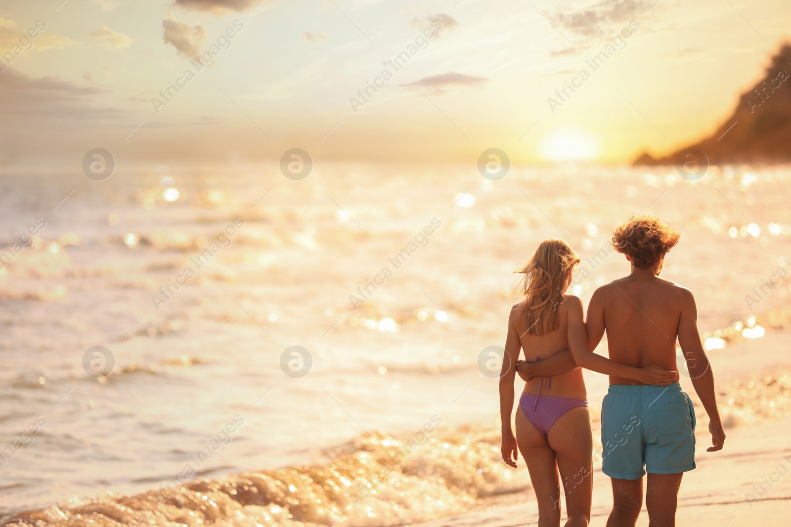 Photo of Young woman in bikini and her boyfriend walking on beach at sunset. Lovely couple