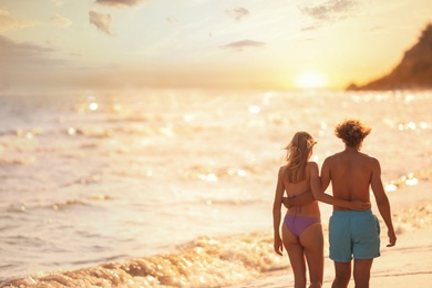 Photo of Young woman in bikini and her boyfriend walking on beach at sunset. Lovely couple