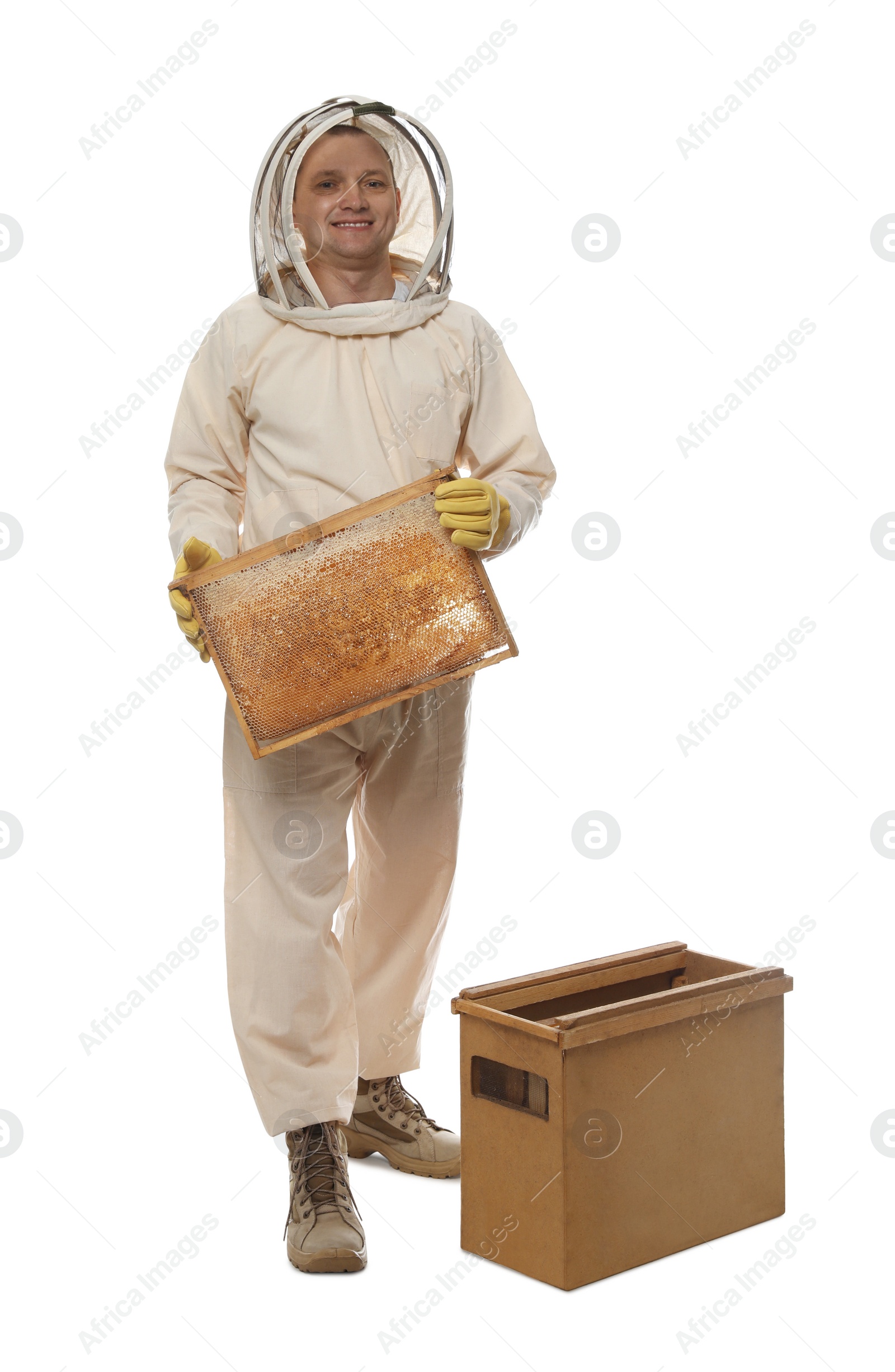 Photo of Beekeeper in uniform holding frame with honeycomb near wooden hive on white background