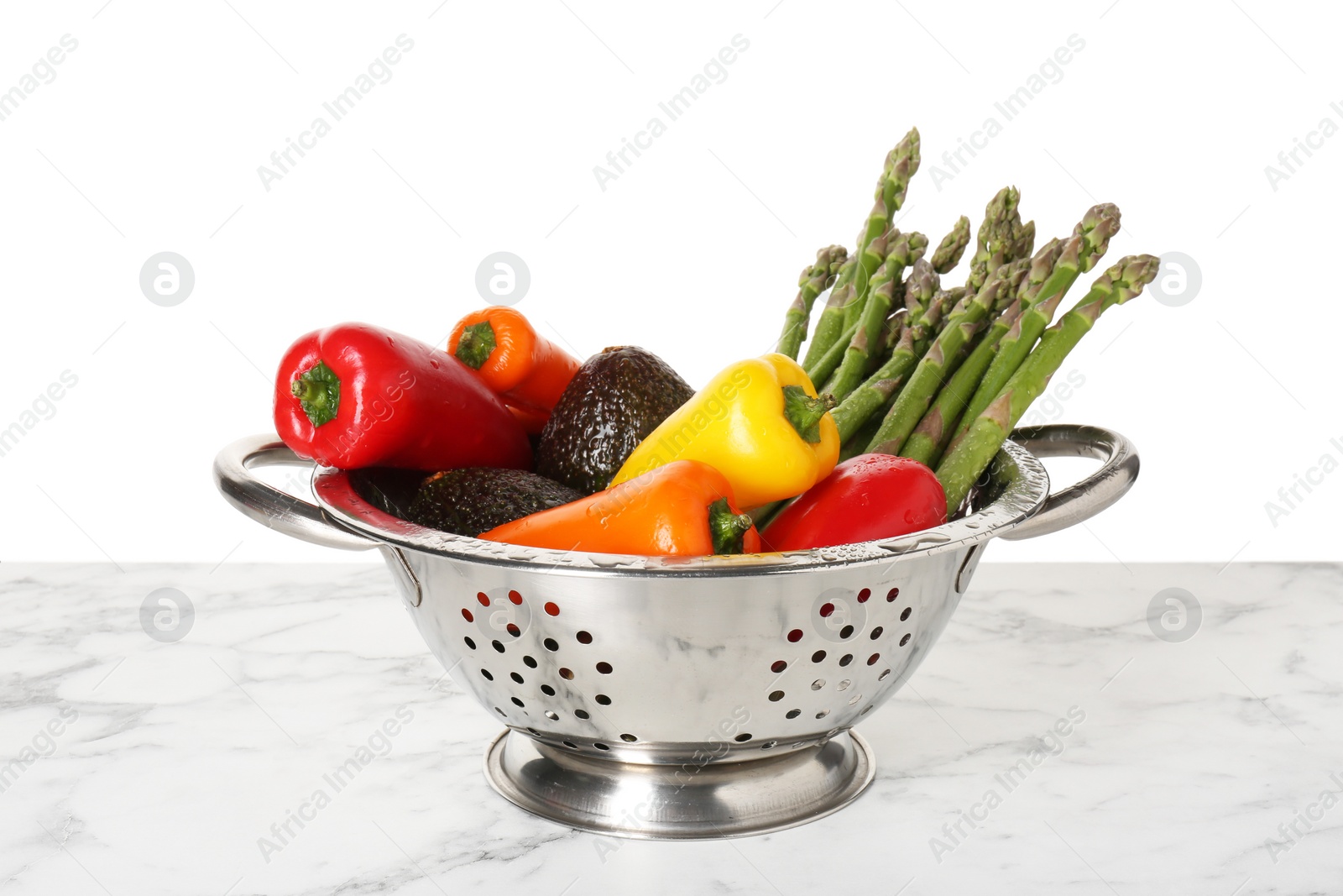 Photo of Metal colander with different vegetables and avocados on marble table against white background