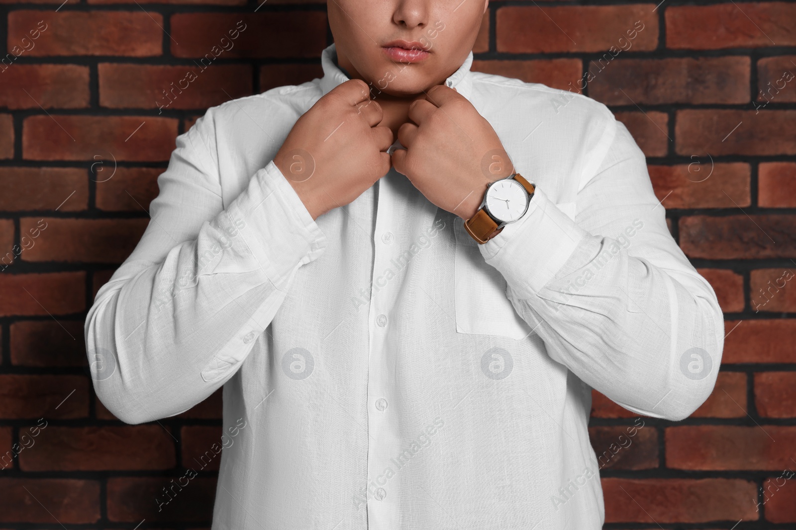 Photo of Groom putting on elegant white shirt near red brick wall, closeup. Wedding day