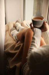 Photo of Woman in knitted socks holding cup of tea on plaid near window at home, closeup