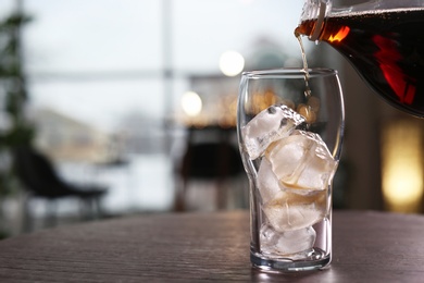Photo of Pouring cola into glass with ice on table against blurred background. Space for text