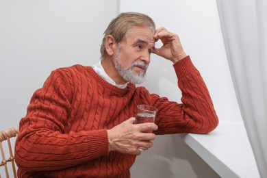 Photo of Upset senior man with glass of water looking at window indoors. Loneliness concept