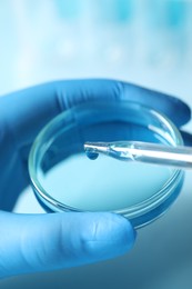 Photo of Doctor dripping liquid from pipette into petri dish on light blue background, closeup. Laboratory analysis